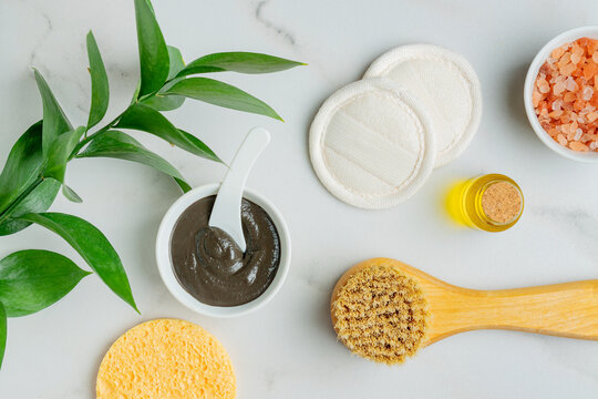 Homemade clay mask for the face and body in a white bowl next to assorted sponges. Reusable lufa pads for mask removing and face prush for dry massaging on a table. Spa procedures at home.