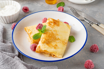 Traditional thin pancakes or crepes with cottage cheese, honey, sour cream and raspberries with cup of tea on a gray concrete background. Food for Maslenitsa. Copy space