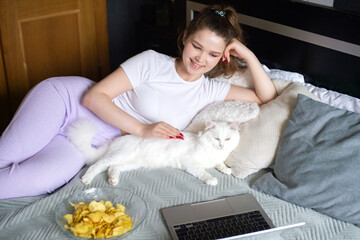 A cute girl is lying on a bed with a cat and looking at a laptop