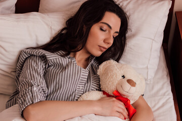A young beautiful girl sleeps with a teddy bear in a bed in pajamas