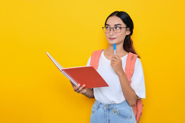 Thoughtful pretty young student girl wearing backpack and eyeglasses holds notebook and pen, looking up at copy space isolated on yellow background