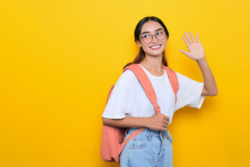 Cheerful pretty young student girl wearing backpack and eyeglasses waving hand say hello isolated on yellow background