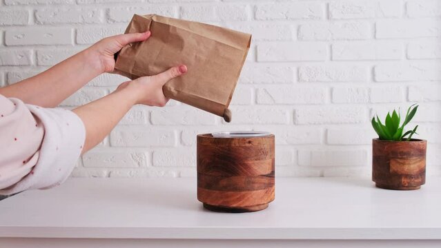 Woman gardener pours drainage for planting a houseplant in a pot, home interior. Female hands pour stone drainage into a flower pot for soil preparation, white brick wall background
