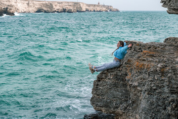 A woman in a blue jacket sits on a rock above a cliff above the sea, looking at the stormy ocean. Girl traveler rests, thinks, dreams, enjoys nature. Peace and calm landscape, windy weather.