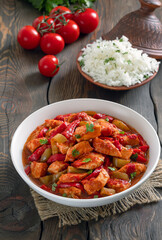 Tomato chicken stew. Braised chicken breast in tomato sauce mixed with onion, red and green bell pepper served with bowl of cooked rice on background. Wooden background, vertical, selective focus.