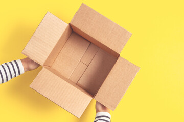 Woman hands holding one empty open brown cardboard box on yellow background. Top view