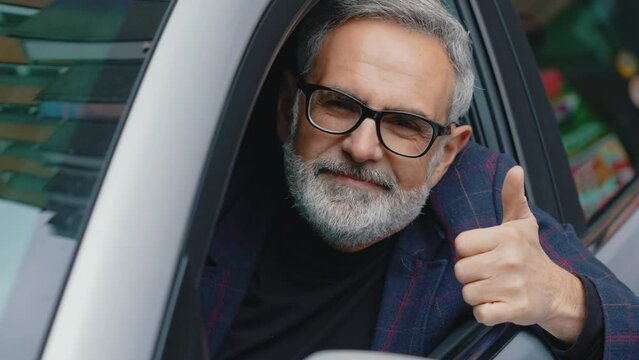Positive driver. Outdoor closeup shot of friendly caucasian gray haired man with gray facial hair and black-frame glasses peeking out of driver's seat window, smiling at camera and showing thumbs up