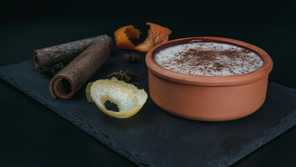 Rice pudding dessert served in an earthenware dish on a slate table accompanied by lemon and orange peel and cinnamon sticks. Foodie