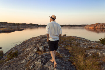 A caucasian man with straw hat and a white linnen shirt walking on a rock by the sea at sunset.