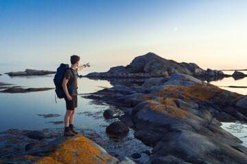 Man hiking by the sea with a backpack at sunset pointing with his arm.