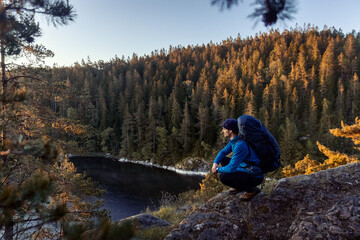 A caucasian man with a backpack sitting on a rock in a forest next to a lake at sunrise.