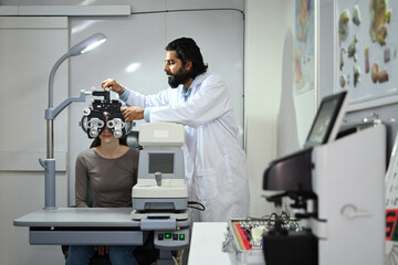 Male ophthalmologist with beard in white examining checking eye vision of patient with phoropter eyesight measurement testing machine