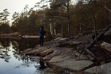 A caucasian man walking on a rock with a backpack by a lake in a forest at sunrise.