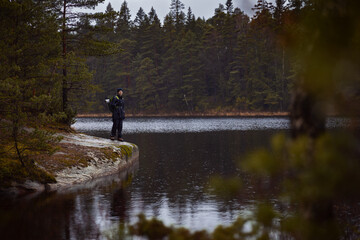A caucasian man with a backpack standing on a rock by a lake in a forest on a rainy day.
