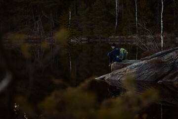 A caucasian man sitting on a rock with a backpack by a lake in a forest.