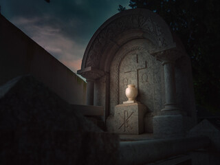 Fototapeta premium Low light dramatic and smooth red tinted tomb in a obscure Christian cemetery adorned with a carved floral design with an urn illuminated by a beam of light at night in Vandellòs, Catalonia, Spain