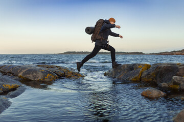 A caucasian man jumping over a gap on a rock by the sea with a backpack.