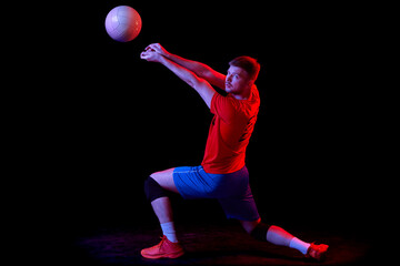 Dynamic portrait of male volleyball player training with ball isolated on dark background in neon...