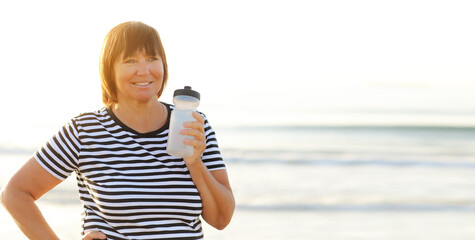 mature woman drinking water from bottle after fitness exercises or yoga on beach by sea. fitness and healthy lifestyle concept - thirsty senior woman with mat maintains water balance after sport