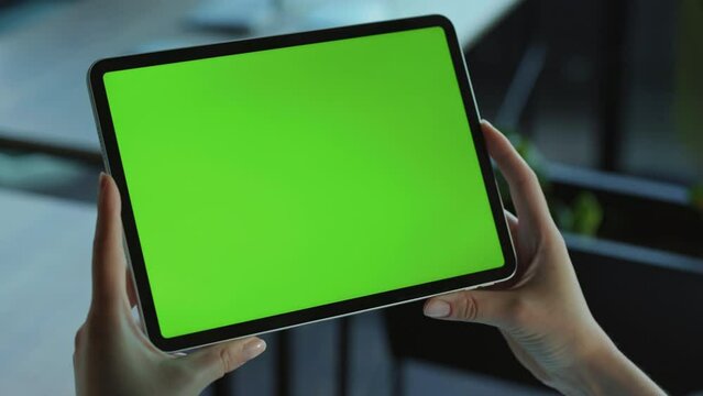 Hands of female office worker holding tablet with green screen. Horizontal view of gadget. Caucasian young woman browsing. Touchscreen. Modern technologies. Blurred background. People, person