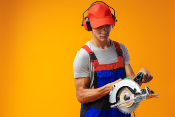 Young male construction worker with modern circular saw on yellow studio background