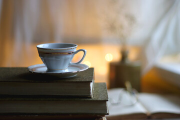 Plate of chocolate chip cookies, stack of vintage books, reading glasses, cup of tea or coffee, lit candle and fairy lights. Hygge at home. Selective focus.