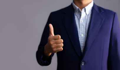 Businessman in dark blue suit hand thump up closeup grey background.
