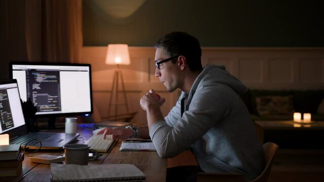 young involved man in glasses remote works at home on laptop at night. bites apple, types fast focusing on monitor and keyboard. hardworking and concentrated on programming job with apple in mouth