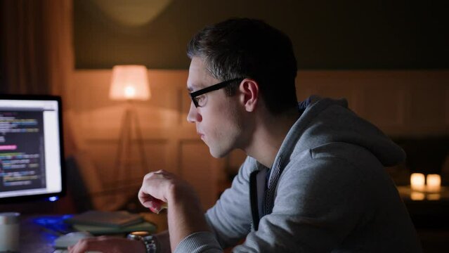 young man in glasses remote works at home, sits at table with monitor and laptop at night. bites apple, types on keyboard looking intently on screen. hardworking and concentrate on programming job
