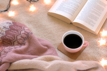 Concept of relaxation and hygge on a winter day. Top view of a cup of black coffee and an open book on the bed with Christmas lights. Copy space.