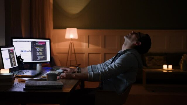 young overworked freelancer leans back on chair, throws back head, falls asleep, sits at desk with computer at home after working night. workaholic programmer. tired student after exam preparation