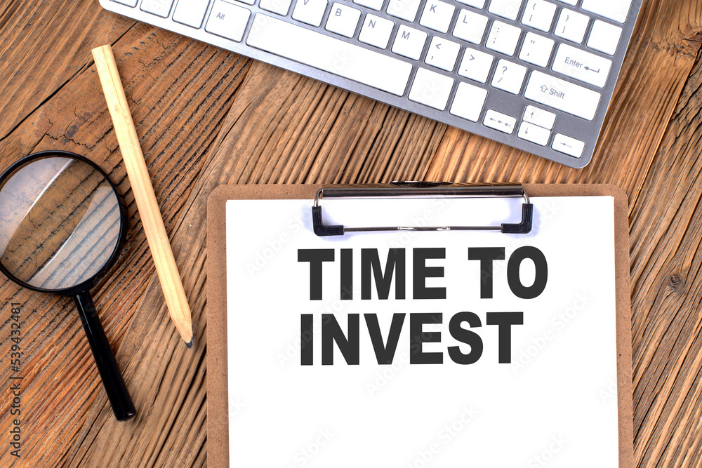 Poster time to invest text on paper clipboard with magnifier and keyboard on wooden background