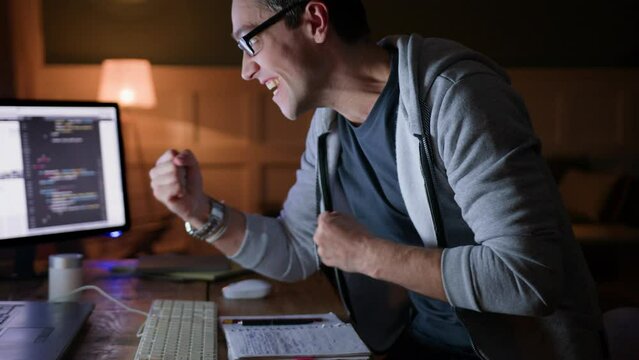 young man remote works or distance studies in home office sitting at table with computer laptop at darkness. gets excited of great news, stands up, screams yeah and laughs, gestures emotionally 