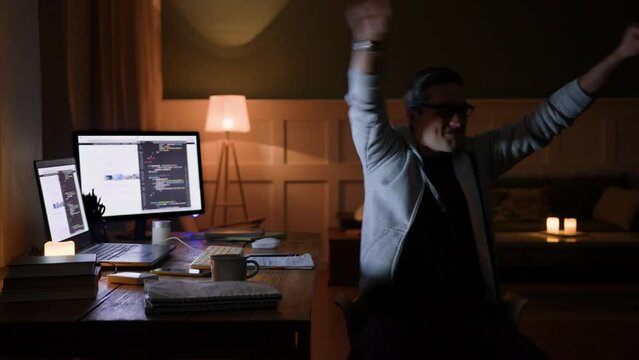 young man remote works sitting at table with computer laptop in darkness. waits nervously, gets excited of great news, spins around on chair, screams yeah and laughs, gestures emotionally 