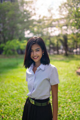 Portrait photo of a young asian beautiful lady in thai university student uniform smile happily in a park with nice sunlight from the back