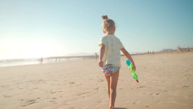 Child playing ocean beach summer day with water gun. Little girl caucasian have fun run on sand seashore with squirt gun. Adorable female 5 years old enjoy playing on beach Handheld. 