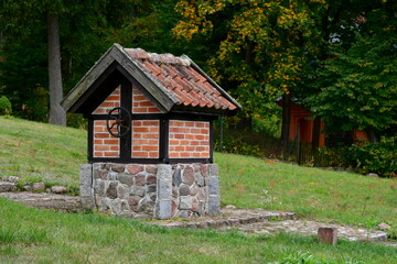 Fototapeta na wymiar A close up on a small well with a metal wheel made out of stones, boulders, red bricks, and tiles, located next to some stone steps, old manor and a church spotted on a gloomy autumn day in Poland