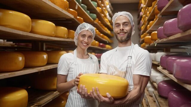 Close up portrait of Caucasian young joyful family couple woman and man smiling to camera standing in cheese storage holding in hands cheese wheel. Dairy products Producing cheese at farm house