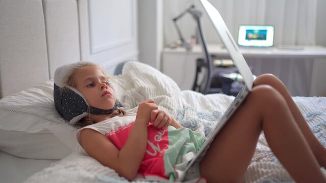 Child in funny hat watching video on laptop lies sofa at home. Little caucasian girl enjoy cartoons on laptop sitting bed in bedroom. Joyful preschool kid girl use laptop computer