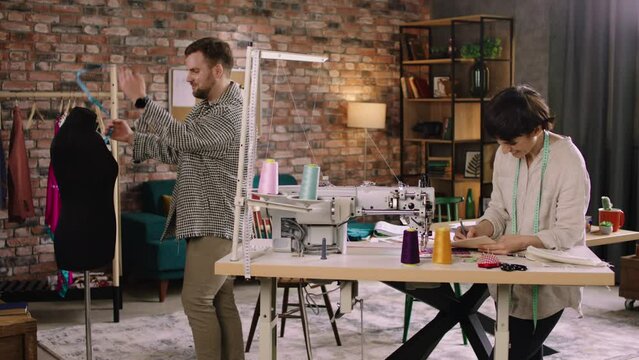 A man is doing up measurements on a mannequin as the woman is doing work at her sewing machine, they both look very pleased and are chatting with eachother