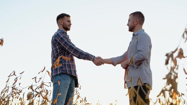 Two farmers standing outdoors in soy field in autumn shaking hands on deal. Handshake on soybean field