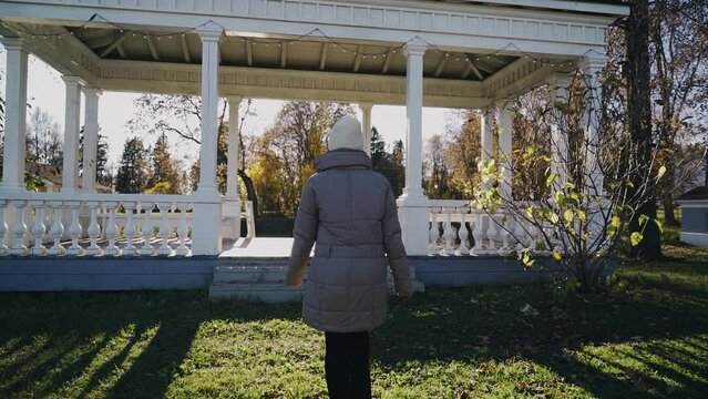 In the direction of the gazebo there is a girl wearing a gray jacket and a hat. Shooting a girl from behind in motion