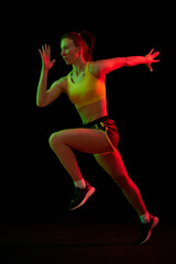 Fototapeta na wymiar Dynamic portrait of young sportive woman training, running isolated on dark background in neon light. Competition, sport, fitness, speed and energy concept.