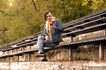 Fototapeta premium Young man with dreadlocks browsing mobile phone, sitting on old shabby bench of street bleacher.