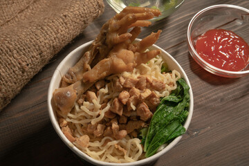 Chicken noodles in white bowl and wooden chopsticks on natural background with chicken pieces, chicken feet, green mustard, sauce. Indonesian Chicken Noodles