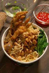 Chicken noodles in white bowl and wooden chopsticks on natural background with chicken pieces, chicken feet, green mustard, sauce. Indonesian Chicken Noodles