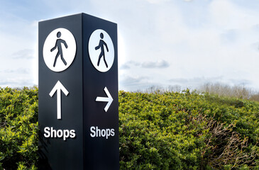 Black and white freestanding directional sign post with arrows and person symbol icon pointing towards shops. 