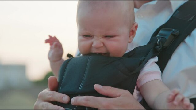 At sunlight cute face portrait of a newborn baby looking around in a slings carrier at sunlight. Young father holding a child on street. Ergo backpack. Parenthood. Dad. City. Slow motion