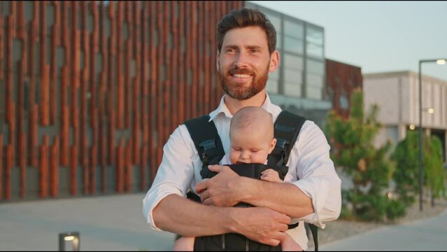 Portrait of a happy young father holding a funny newborn baby in a slings carrier on street. Ergo backpack. Parents. Modern city. Slow motion
