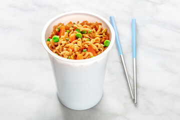 Ramen cup, instant soba noodles in a plastic cup with vegetables, green peas and carrots, with chopsticks, on a white marble background